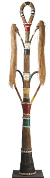Fotografia de uma escultura de formato alongado, com a forma de uma espécie de tronco vertical, com alças e uma forma elíptica no topo. A escultura é decorada com búzios e faixas de couro pintadas com diversas cores, além de fibras de palha.