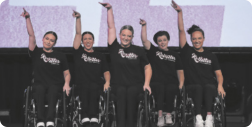 Fotografia de cinco mulheres em cadeiras de rodas em um palco, enfileiradas e uniformizadas, sorrindo com um dos braços erguido para o alto.