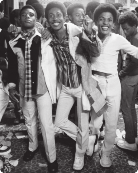 Fotografia em preto e branco de um grupo de pessoas negras com cabelos cacheados com penteado 'black power', usando roupas brancas e coloridas, posando para a fotografia.