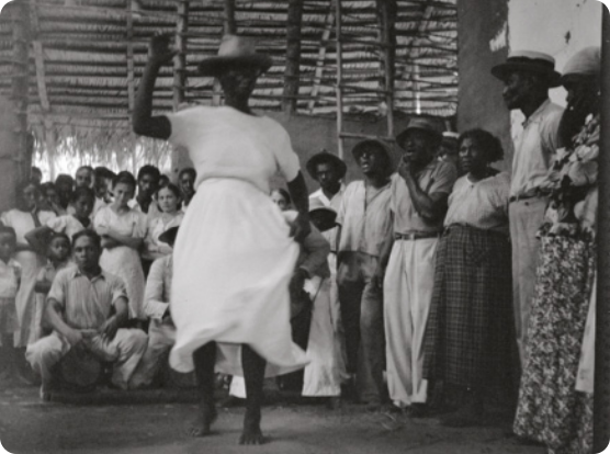 Fotografia em preto e branco de um grupo de pessoas em volta de uma mulher de vestido branco e chapéu e chapéu dançando, descalça, sobre o piso de um terreiro.
