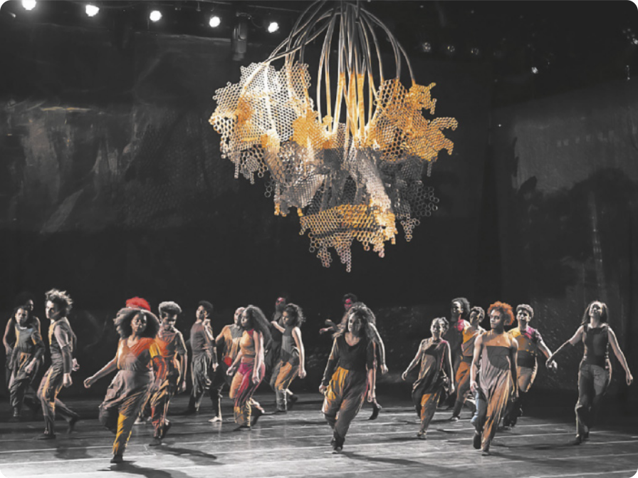 Fotografia de um grupo de artistas dançando em um palco, executando um passo em coreografia.