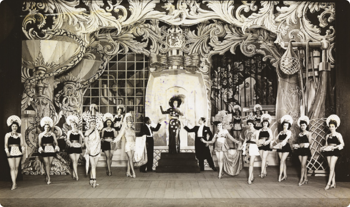 Fotografia em preto e branco de um grupo de dançarinas em um palco, enfileiradas, usando saias curtas e adornos na cabeça, exibindo pequenos aventais na região da cintura. Ao centro está uma mulher de vestido descendo uma escada, usando um chapéu com plumas, sendo recebida por dois homens de fraque, que lhe oferecem as mãos. O cenário ao fundo é complexo e rebuscado, reunindo diversos elementos figurativos e ornamentos.