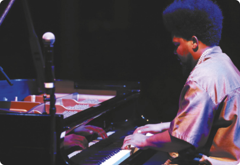 Fotografia de Amaro Freitas. Ele é um homem com barba e cabelos cacheados, usando uma camisa e colares, tocando piano em um palco.