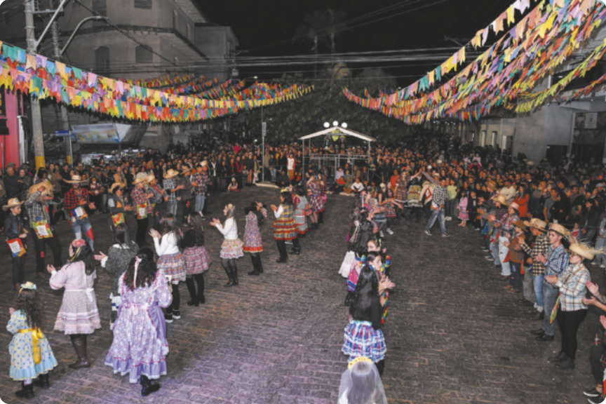 Fotografia de um grupo de jovens e crianças enfileirados frente a frente em uma praça, dançando uma quadrilha em uma festa junina. Há duas fileiras de meninas usando vestidos e saias coloridas, ao centro, e duas fileiras de meninos nas laterais usando camisas xadrez e chapéus de palha. Ao fundo há uma multidão de pessoas assistindo à dança, e a praça está coberta por bandeiras coloridas de festa junina, penduradas em cordões no alto.