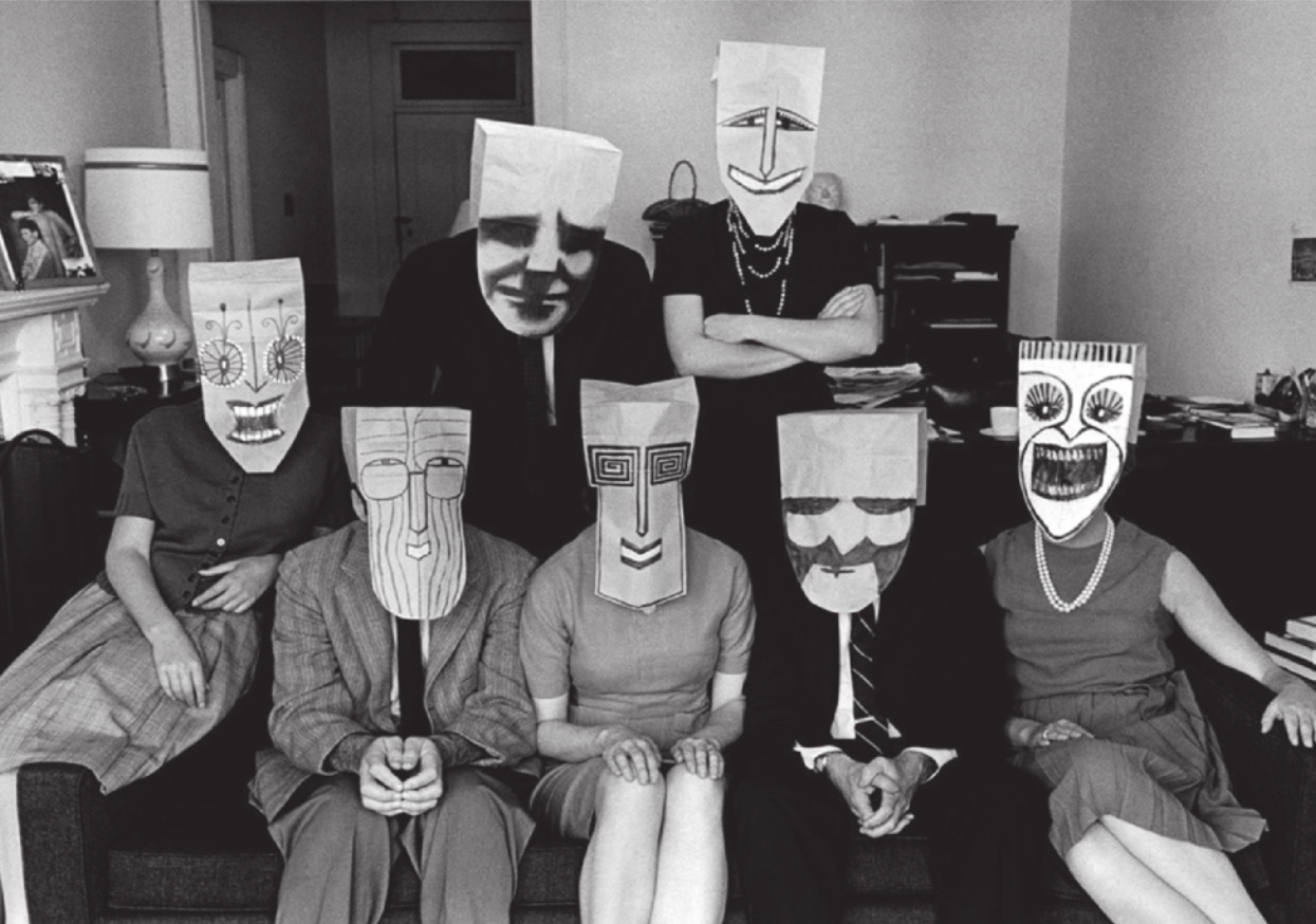 Fotografia em preto e branco de um grupo de pessoas em uma sala, sentadas em um sofá, com dois deles em pé atrás do sofá. Eles estão usando diversas máscaras de papel, cada uma com um formato e um desenho diferente de um rosto cômico e estilizado.