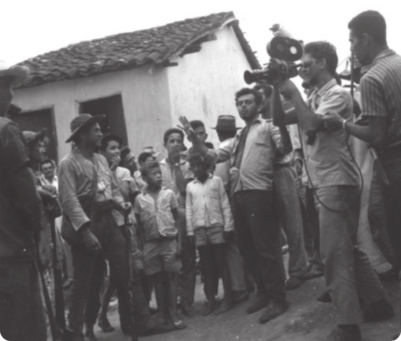 Fotografia em preto e branco de um grupo de pessoas em uma paisagem semiárida, com uma pequena casa ao fundo, em torno de Glauber Rocha. Ele é um homem de barba e cabelos curtos, usando uma camisa, gesticulando com as mãos enquanto fala com o grupo. Ao lado dele há um cinegrafista segurando uma câmera.