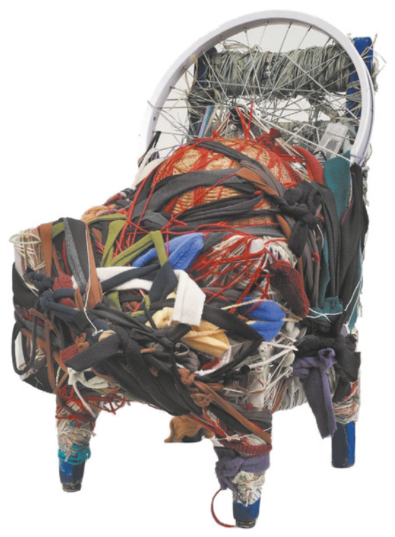 Fotografia de uma escultura feita com uma cadeira completamente revestida com muitos pedaços, tiras e fragmentos de tecidos de diferentes materiais e cores diversas. No encosto da cadeira está também uma roda de bicicleta.