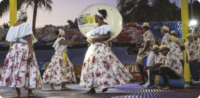 Fotografia de um grupo de mulheres dançando ao ar livre, com saias de estampas floridas e lenços na cabeça. Ao lado delas, há um grupo de homens de chapéu sentados sobre tambores, tocando-os, e homens em pé cantando com microfones.