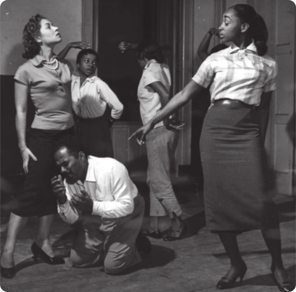 Fotografia em preto e branco de um grupo de atrizes em pé em um cenário teatral, uma delas apontando o dedo indicador para um ator que está ajoelhado no chão, com expressão de tristeza, levando as mãos em direção ao rosto.