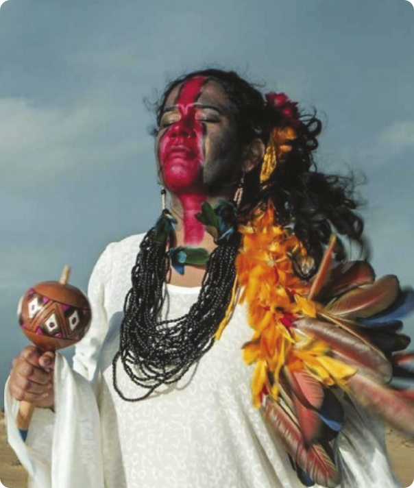 Fotografia de Shirley Djukurnã Krenak. Ela é uma mulher indígena com o rosto pintado e usa colares e enfeites com penas no cabelo. Ela está segurando um chocalho, de olhos fechados, sob um céu nublado.