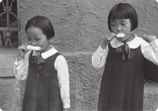 Fotografia em preto e branco de duas meninas de vestido, com cabelos cortados à altura das orelhas, chupando picolés.