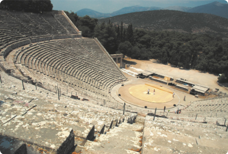 Fotografia de ruínas de um teatro grego, com uma arena central em formato semicircular, um palco retangular atrás da arena e uma grande arquibancada semicircular voltada para o palco. Atrás do palco há uma paisagem com árvores e montanhas.
