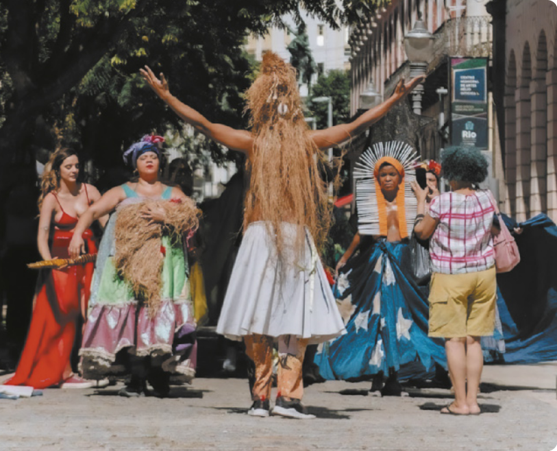 Fotografia de um grupo de artistas em uma calçada, usando trajes de inspiração indígena e afrodescendente. Há uma mulher de vestido com uma espécie de chocalho indígena em forma de bastão; uma mulher de vestido e lenço na cabeça dançando com um pacote de fibras de palha no colo; um homem de saia, de costas, com os braços erguidos para o alto, usando uma espécie de peruca de cabelos longos feitos com palha; e uma mulher vestida com uma saia estrelada e um cocar. Ao lado do grupo há uma mulher filmando a apresentação com um smartphone.
