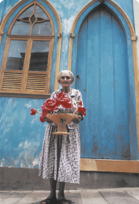 Fotografia de uma mulher idosa de vestido, segurando um vaso com grandes flores vermelhas artificiais. Ela está em pé em uma calçada em frente a uma casa azul, com a porta e a janela em forma de arcos.