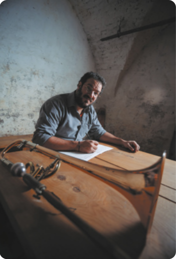 Fotografia de Jonas Hocherman. Ele é um homem com barba e óculos, escrevendo em uma folha de papel sobre uma mesa, na qual está apoiado um trombone.