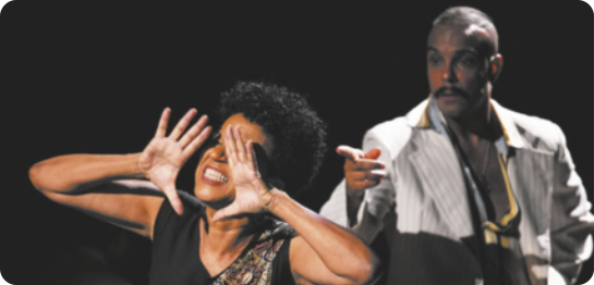 Fotografia de atores em um palco. À frente está Juçara Marçal: uma mulher com cabelos curtos e cacheados, com as mãos abertas à frente do rosto; atrás dela, Jé Oliveira, um homem com bigode e cabelo raspado, usando um paletó, aponta o dedo indicador para ela.