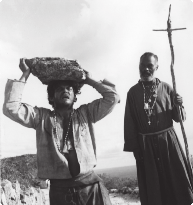 Fotografia em preto e branco de um homem de camisa aberta carregando uma pedra sobre a cabeça, com expressão de sofrimento. Atrás dele está caminhando um homem de barba com diversos colares, vestido com uma batina e carregando um cajado com uma cruz no topo. Eles estão caminhando em uma paisagem semiárida.