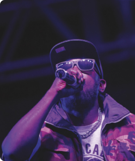 Fotografia de Fernandinho Beat Box. Ele é um homem com barba, de boné, óculos escuros e corrente no pescoço, cantando com um microfone na mão, vestido com uma jaqueta.