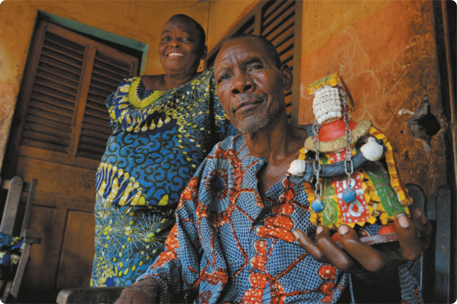 Fotografia de Cyprien Tokoudagba. Ele é um homem idoso com cabelos curtos e barba grisalha, vestido com uma camisa com estampas coloridas. Ele está sentado em uma cadeira, segurando uma escultura que representa uma figura humana sem rosto, com um vestido colorido e correntes pendendo do cabelo, segurando uma espécie de facão. Ao lado dele está uma mulher de cabelos curtos, em pé, com um vestido colorido estampado com formas abstratas.