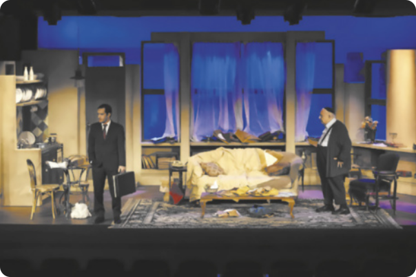 Fotografia de dois homens atuando em um palco, com um cenário representando uma sala de estar com um sofá e uma mesa de centro sobre um tapete. À esquerda está Ricardo Gelli: um homem vestido com terno e gravara, carregando uma maleta; e à direita está Sérgio Mamberti: um homem idoso de terno e suspensórios, usando um quipá na cabeça.