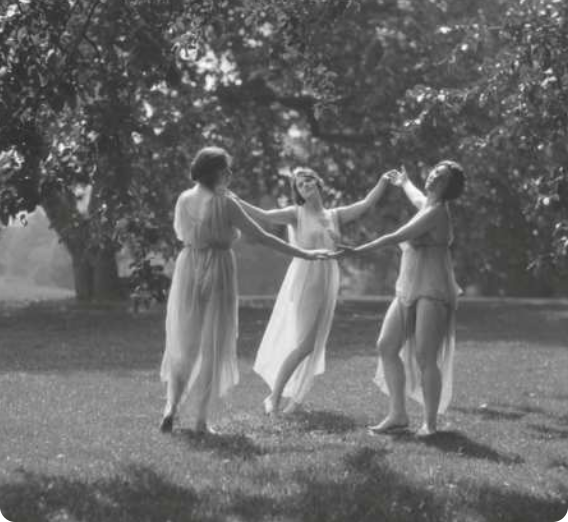 Fotografia em preto e branco de três mulheres de vestido dançando em roda, de mãos dadas, em um gramado, com árvores ao fundo.