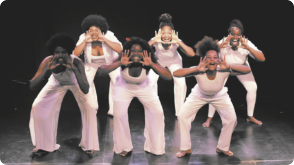 Fotografia de um grupo de mulheres em um palco, uniformizadas com roupas brancas, enfileiradas em duas linhas de três mulheres cada, todas com os joelhos flexionados e o tronco inclinado para a frente, com as mãos abertas ao lado da boca.