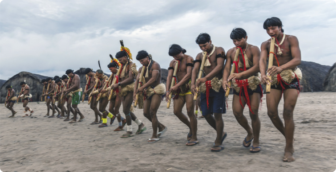 Fotografia de um grupo de homens indígenas dançando enfileirados, tocando flautas, no solo de areia de uma aldeia. Eles estão usando adereços de palha, cocares e outros adornos indígenas.
