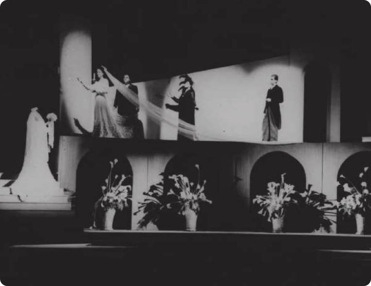 Fotografia em preto e branco de atores em um palco. O cenário tem vasos com plantas, arcos, e uma escada em que está uma mulher de vestido segurando um buquê de flores. No andar de cima do cenário está uma mulher com um vestido de noiva que se dirige à mulher na escada com os braços abertos. Atrás da mulher com vestido de noiva há três atores, usando ternos.