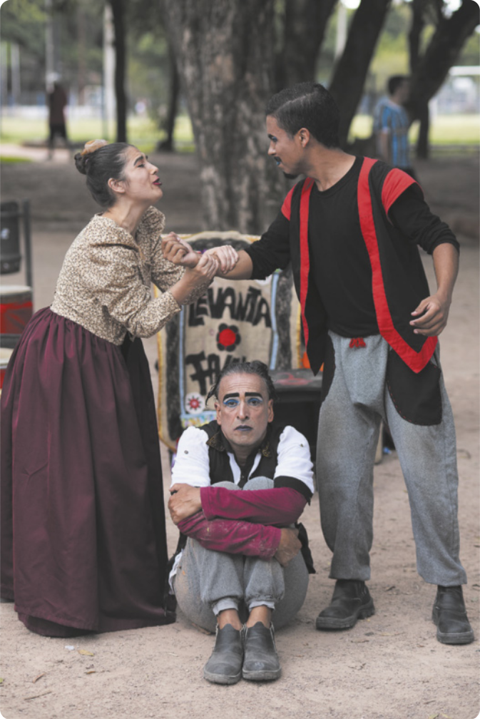 Fotografia de três atores contracenando em uma praça pública, com árvores ao fundo. Uma mulher de saia e cabelos presos em um coque segura o braço de um homem de colete, com expressão de súplica. Entre eles, há um homem de óculos sentado no chão, encolhido, abraçando as pernas com expressão desolada.