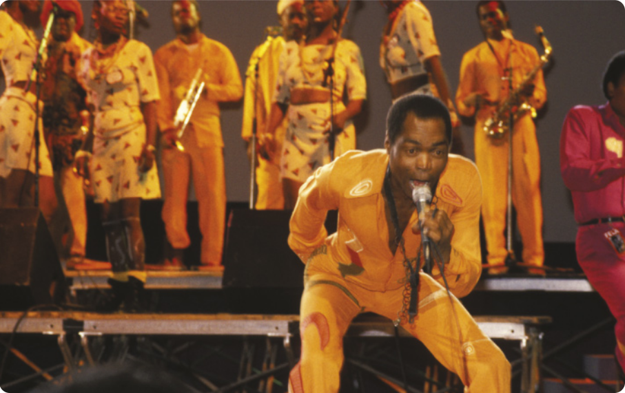 Fotografia de Fela Kuti: um homem com cabelos curtos cantando em um microfone em um palco, com as pernas flexionadas e o tronco inclinado para a frente. Ao fundo, há uma banda com diversos músicos segurando instrumentos de sopro e mulheres cantando.