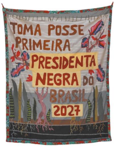 Fotografia de um tecido com bordados representando árvores e plantas, além da frase: 'Toma posse primeira presidenta negra do Brasil: 2027'.