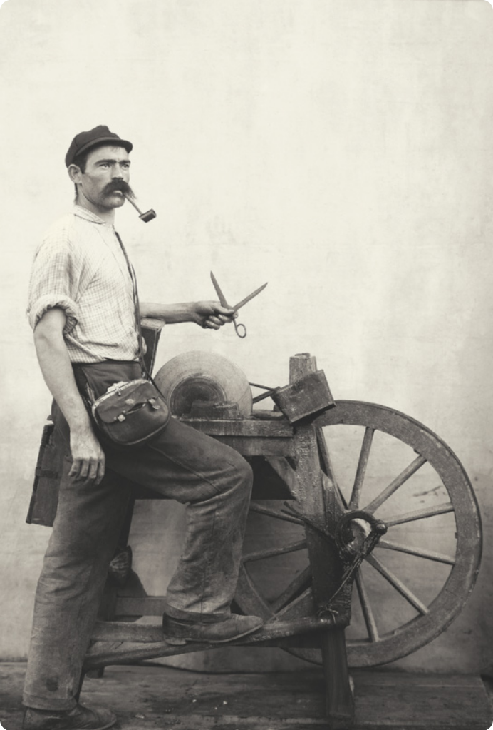 Fotografia em preto e branco de um homem de boné com um bigode longo, um cachimbo na boca e uma tesoura na mão. Ele está com um dos pés apoiado em um pedal, que aciona a roda de um mecanismo de madeira que faz girar uma pedra de formato circular usada para amolar lâminas. A composição da fotografia é bem equilibrada e mostra cada um dos elementos da imagem de maneira clara, com ênfase na roda da máquina de amolar e na tesoura ao centro. A pose do amolador é imponente e altiva, como em uma escultura clássica, e é visível que o homem retratado está posando para a fotografia.