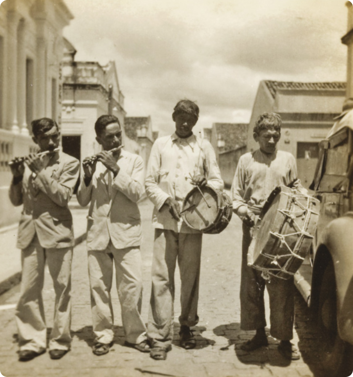 Fotografia em preto e branco de quatro homens caminhando em uma via pública com piso de pedras e casas ao fundo, tocando instrumentos musicais. À esquerda, dois deles estão tocando pífanos, e os outros dois à direita tocam um tambor pequeno e um tambor grande, respectivamente.