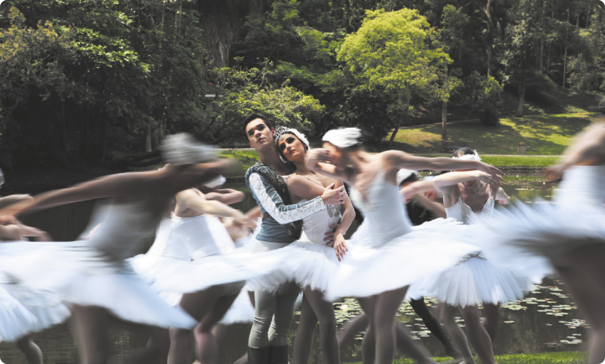 Fotografia de bailarinas vestidas de branco dançando em torno de um casal de bailarinos ao centro, abraçados. Ao fundo há um lago e árvores, em um jardim botânico.
