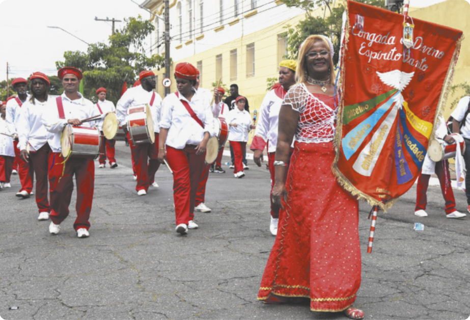 Fotografia de um grupo de pessoas uniformizadas em uma via pública, caminhando em filas e tocando instrumentos de percussão. À frente do grupo está uma mulher de vestido carregando um estandarte com a figura de uma pomba branca, em que está escrito 'Congada do Divino Espírito Santo'.