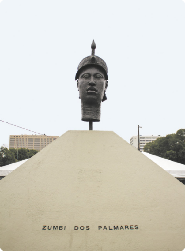 Fotografia de monumento com uma escultura no topo representando a cabeça de um homem usando um capacete com uma espécie de ponta de flecha no topo. No pedestal sob a escultura está escrito 'Zumbi dos Palmares'.