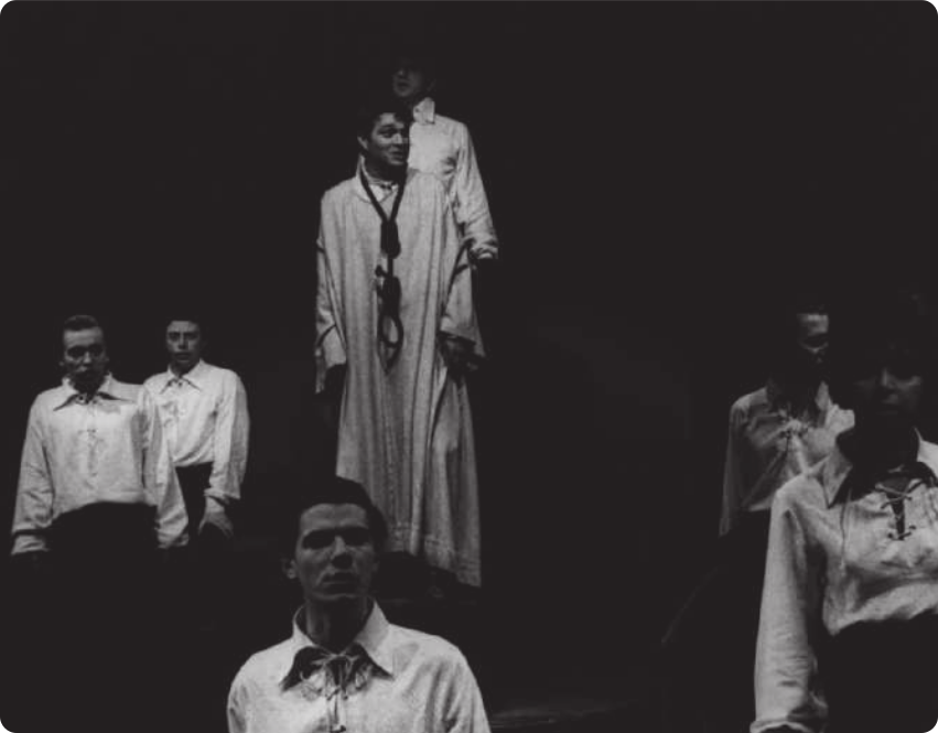 Fotografia em preto e branco de atores em um palco. Há cinco homens, entre eles Gianfrancesco Guarnieri, e uma mulher. Todos estão vestidos com camisas brancas, em pé, com os braços junto ao corpo; e ao centro está David José: um homem com uma espécie de túnica e uma corda no pescoço.