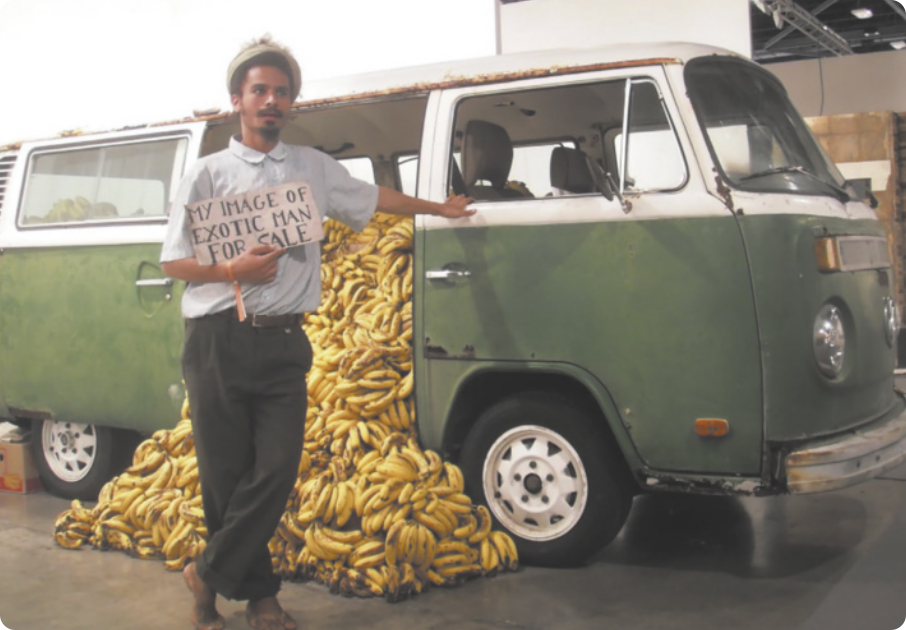 Fotografia de Paulo Nazareth. Ele é um homem com barba e bigode, descalço, usando um chapéu, e está realizando uma performance em frente a uma Kombi com a porta aberta, de onde caem centenas de cachos de banana. Ele carrega uma placa com as palavras, em inglês: 'My image of exotic man for sale'.