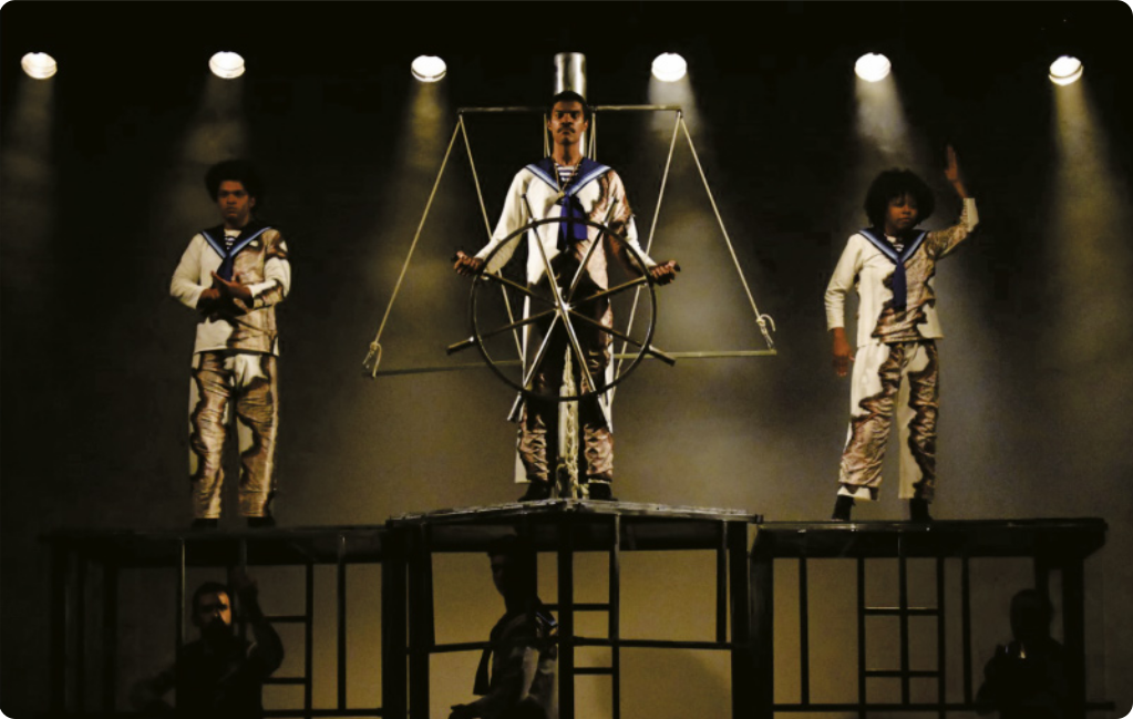 Fotografia de três artistas em um palco vestidos com trajes de marinheiro. Eles estão em pé sobre plataformas elevadas em estruturas metálicas, e um deles está ao centro, segurando um timão de navio.