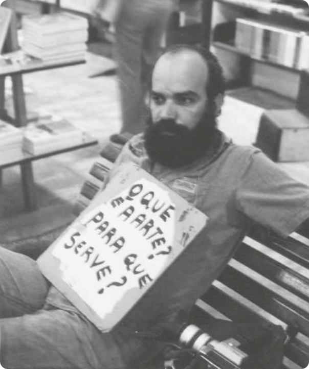 Fotografia em preto e branco de Paulo Bruscky sentado em um banco. Ele é um homem calvo com a barba cheia e escura, usando uma camisa e uma placa em que se lê: 'O que é a arte? Para que se serve?'