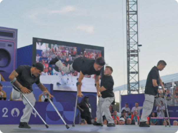 Fotografia de um grupo de 4 atletas paralímpicos em um palco, executando movimentos de breaking com o auxílio de muletas.