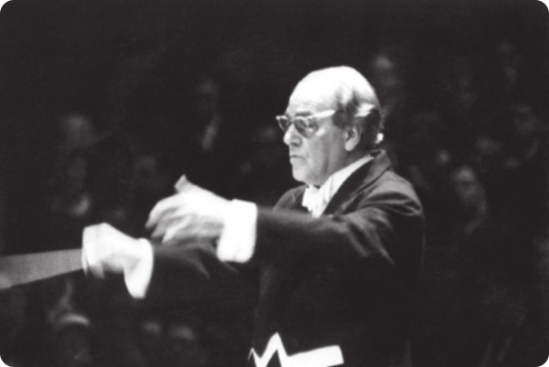 Fotografia em preto e branco de Heitor Villa-Lobos: um homem calvo, de óculos, usando fraque, regendo uma orquestra com uma batuta na mão.