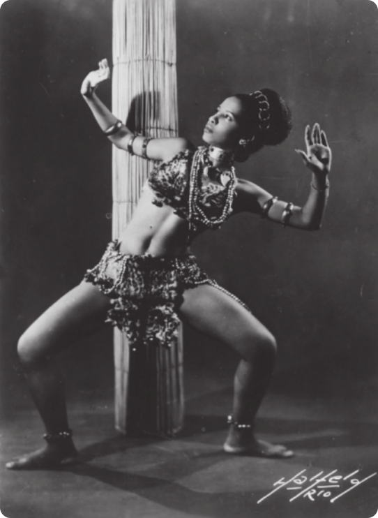 Fotografia em preto e branco de Mercedes Baptista. Ela é uma dançarina vestida com uma saia e um sutiã enfeitados com adereços e adornos diversos, inspirados em trajes de culturas africanas. Ela está com as pernas afastadas e os joelhos flexionados, erguendo os braços ao lado da cabeça e olhando para o alto, com o corpo inclinado para o lado. Atrás dela há um grande objeto cilíndrico feito de fibras de palha.