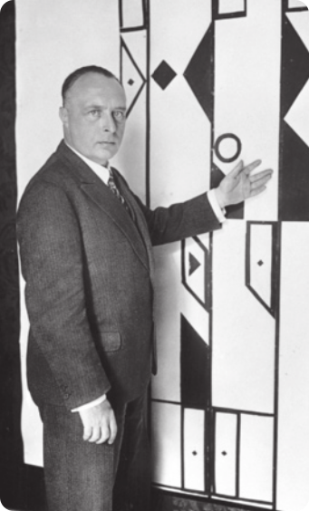 Fotografia em preto e branco de Rudolf Laban. Ele é um homem calvo, com cabelos curtos, vestido com terno e gravata. Ele está em pé, indicando com a mão aberta um desenho geométrico abstrato em uma parede.