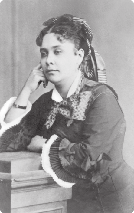 Fotografia em preto e branco de Chiquinha Gonzaga. Ela é uma mulher de cabelos presos, com um lenço na cabeça, usando brincos e um vestido com um laço em volta do pescoço, com os braços apoiados em um móvel e uma das apoiando a lateral do rosto.