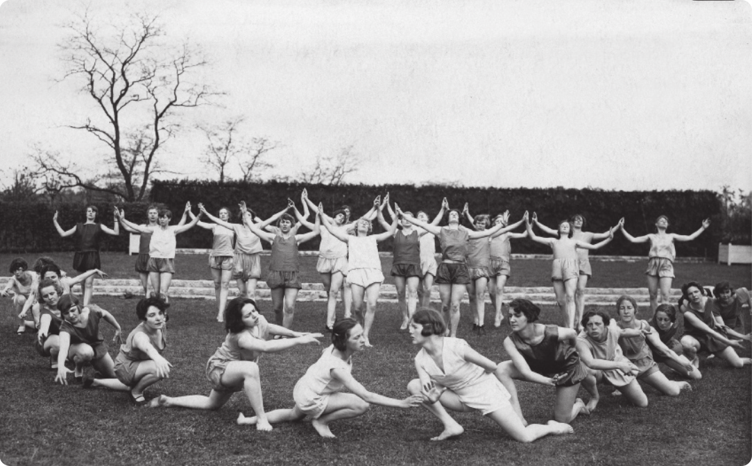 Fotografia em preto e branco de um grupo de mulheres descalças, de bermuda e camiseta, realizando um movimento coreografado em um gramado. Algumas delas estão agachadas, com um dos joelhos apoiado no chão e o tronco inclinado para a frente, em duas filas, formando um semicírculo no chão. Elas estão à frente de outro grupo de mulheres em pé, enfileiradas, com os braços abertos ao lado da cabeça, de mãos dadas, olhando para o alto.