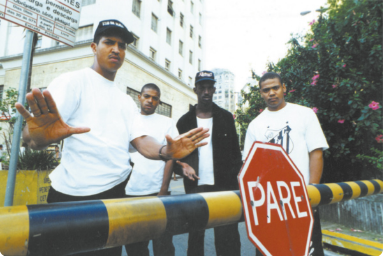 Fotografia de um grupo de quatro rappers do grupo Racionais MC's. Eles estão atrás de uma cancela, com uma placa que diz 'pare' sobre ela. Dois deles estão de boné: um deles é Mano Brown, e ele está mostrando as palmas das mãos abertas em frente ao corpo, gesticulando de acordo com a palavra escrita na placa.