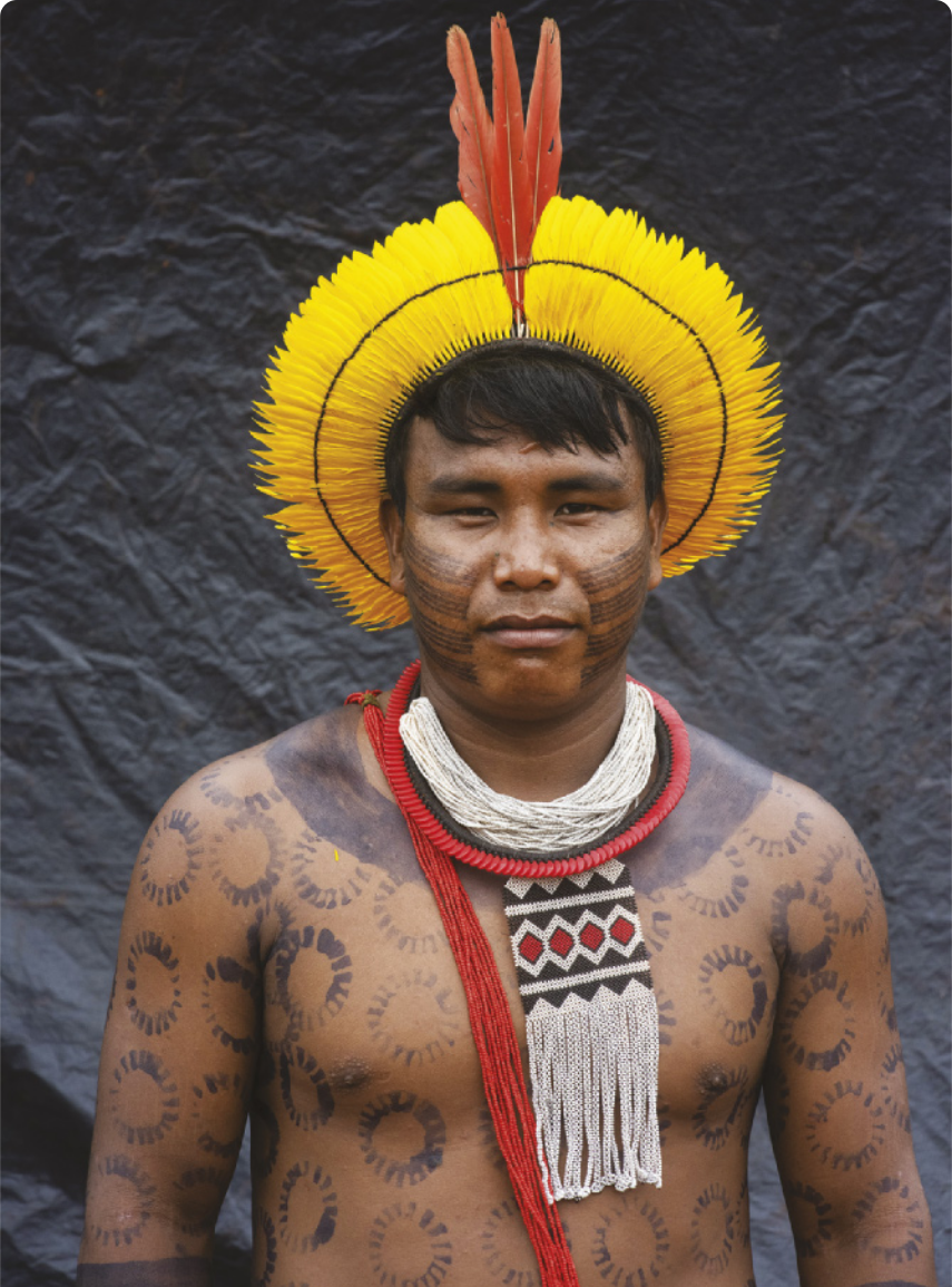 Fotografia de um homem indígena com pinturas corporais de formato circular e linhas horizontais pintadas nas bochechas. Ele usa colares e um cocar de penas.