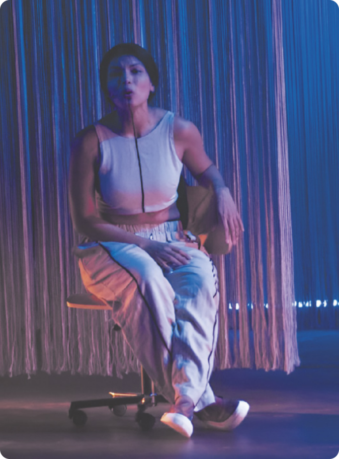 Fotografia de Zahy Tentehar. Ela é uma mulher indígena e está sentada em uma cadeira em um palco, com uma linha vertical pintada no centro do queixo, descendo em direção ao pescoço.
