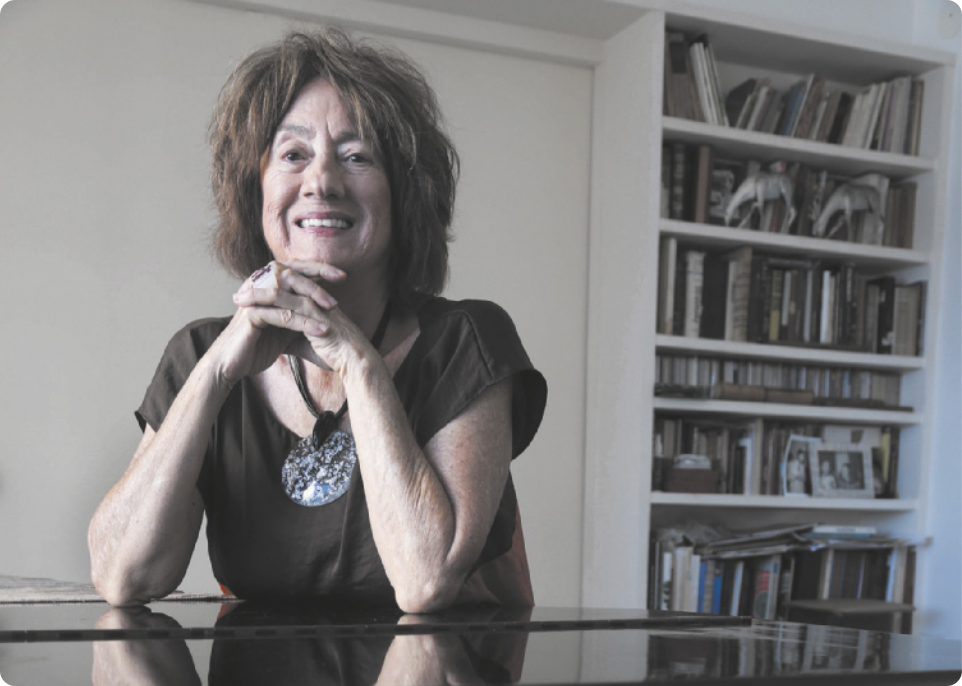 Fotografia de Jocy de Oliveira. Ela é uma mulher idosa com cabelos longos, usando um colar. Ela está sentada diante de uma mesa, sorrindo, com as mãos unidas e os cotovelos apoiados na mesa.